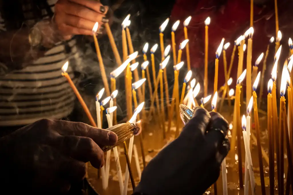 Hands lighting slender beeswax candles among rows of others already burning.